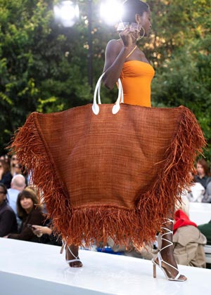 black model on runway carrying enormous straw bag with raw hems and white leather handles detailing in burnt orange colour