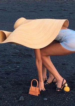 model in enormous straw hat posing with orange microbag on the black sand beach
