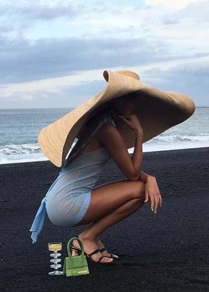 model in enormous straw hat posing with orange microbag on the black sand beach